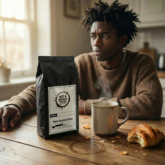 Man sitting at a table looking like "I dont want to be awake right now".  On the table sits a  not a morning person coffee co bag, a stained coffee cup, a half eaten croissant, and a coffee stain.